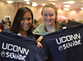 Two students smiling while holding blue UConn Senior T-shirts